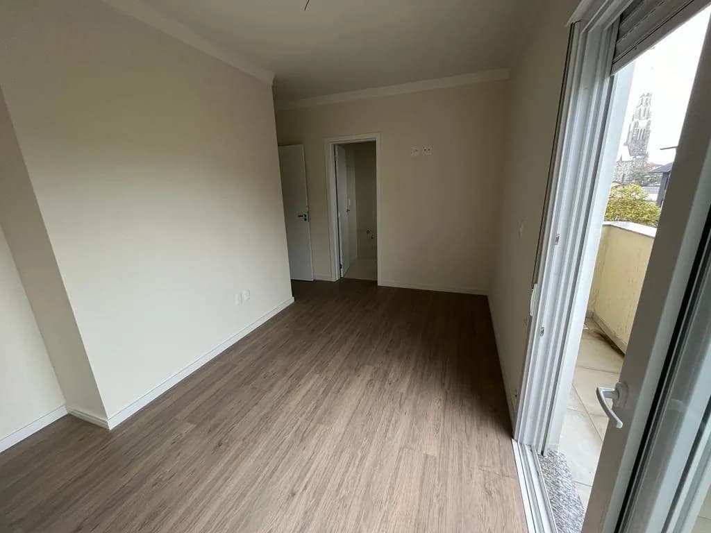 Quarto com piso laminado claro, porta ao fundo e porta de vidro aberta para a sacada, com a torre da Catedral de Pedra ao longe