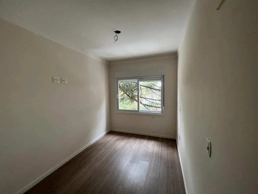 Quarto com piso laminado, paredes claras e janela de correr com persiana voltada para uma araucária