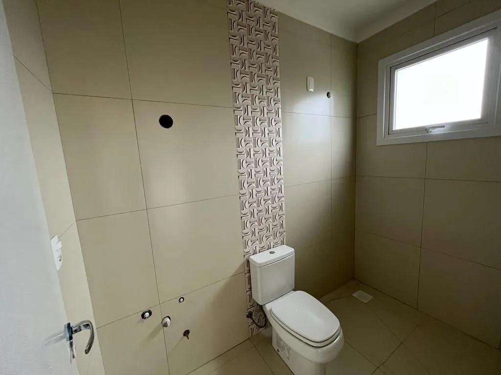 Banheiro com porcelanato bege, faixa decorativa vertical, vaso sanitário, janela e pontos hidráulicos aparentes