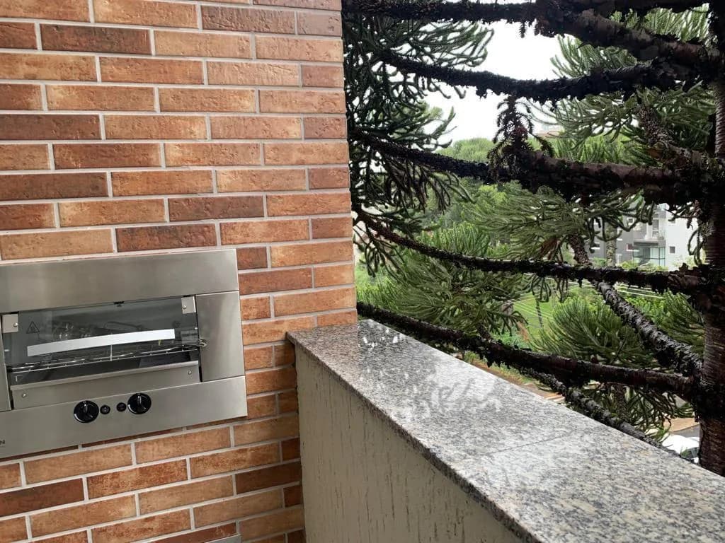 Churrasqueira de inox com vidro embutida na parede de tijolinho da sacada, ao lado do parapeito de granito e de uma araucária