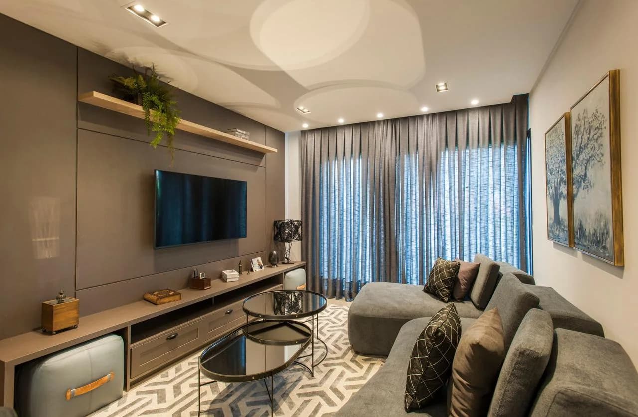 Sala de estar com sofá cinza em L, mesas de centro de vidro, painel de TV com prateleira de madeira e cortinas cinza do teto ao chão