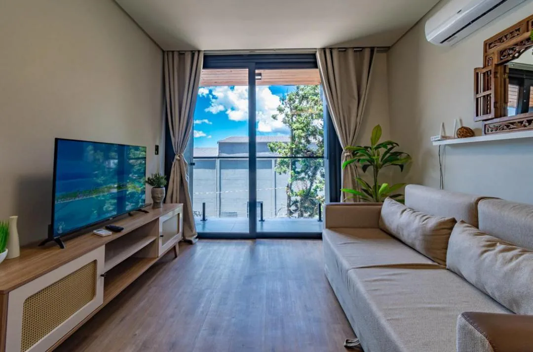 Sala com sofá bege, TV sobre rack de madeira com portas de palhinha e porta de vidro para a sacada