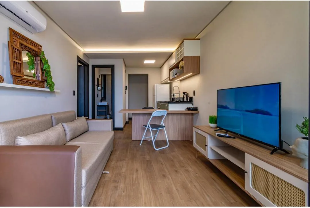 Sala integrada à cozinha, com sofá bege, TV no rack de palhinha e bancada de refeições ao fundo