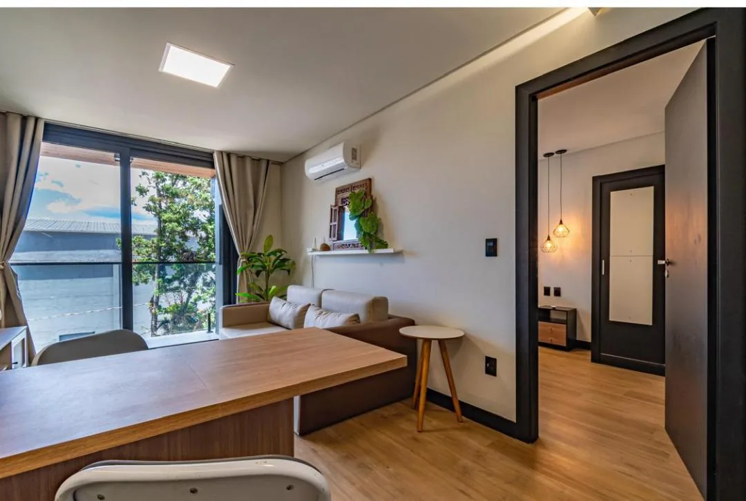 Mesa de refeições em madeira, sofá junto à janela e porta aberta para o quarto com pendentes acesos