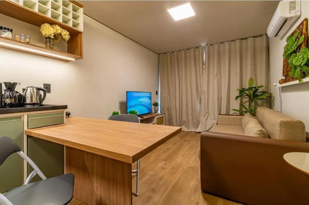 Mesa de madeira encostada na bancada da cozinha, com sofá, TV e cortinas bege ao fundo
