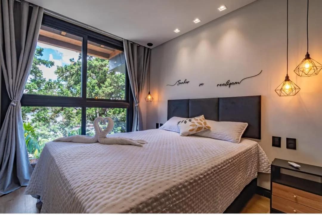 Quarto de casal com janela ampla voltada para árvores, cabeceira estofada escura e pendentes aramados