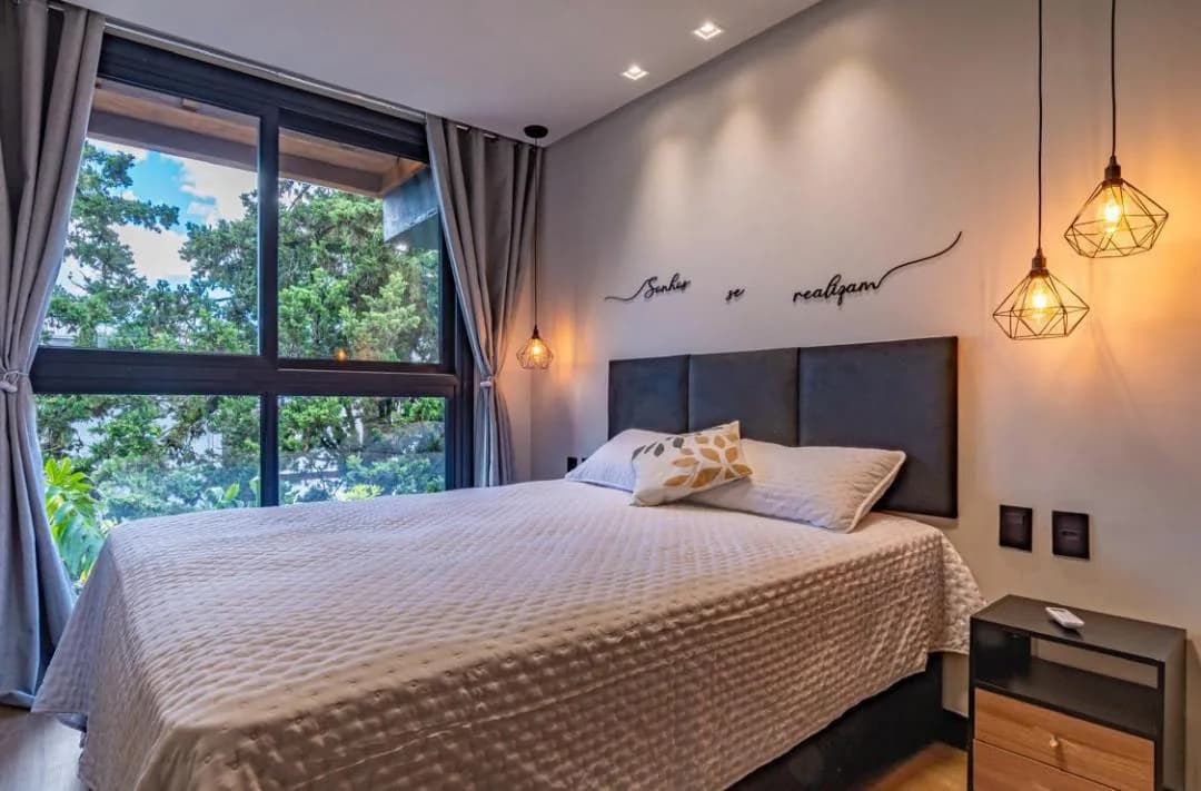 Cama de casal com colcha cinza, criado-mudo e janela ampla com vista para árvores