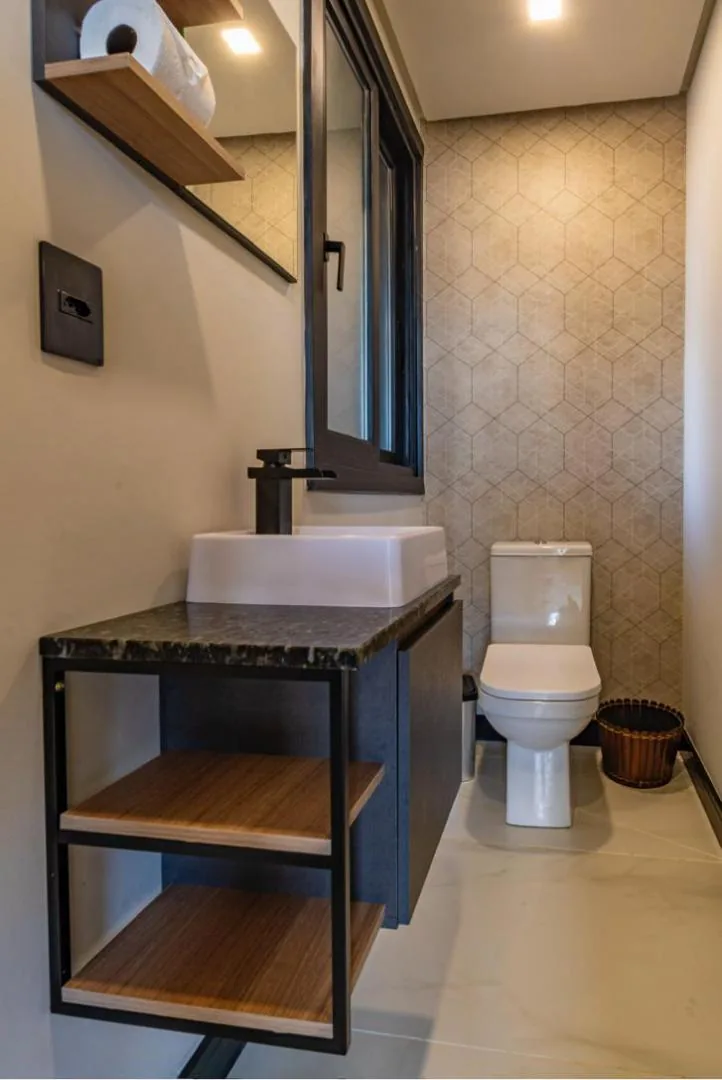 Lavabo com cuba branca sobre bancada de granito, estante metálica com prateleiras de madeira e parede de revestimento geométrico