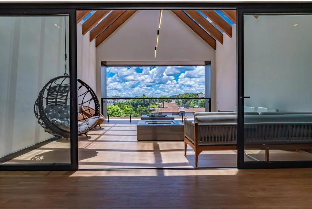Lounge do terraço visto pela porta de vidro, com poltrona suspensa, sofás e grande abertura com guarda-corpo de vidro e vista para a cidade