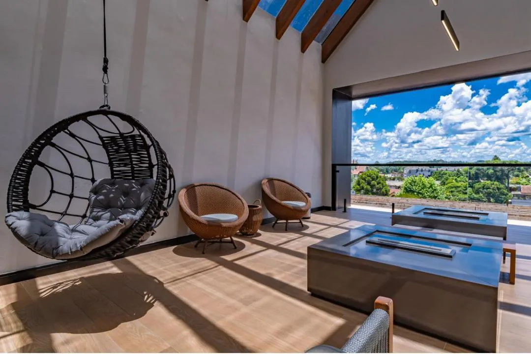 Lounge do terraço com poltrona suspensa, poltronas de fibra e mesas baixas escuras diante de uma grande abertura com vista para a cidade