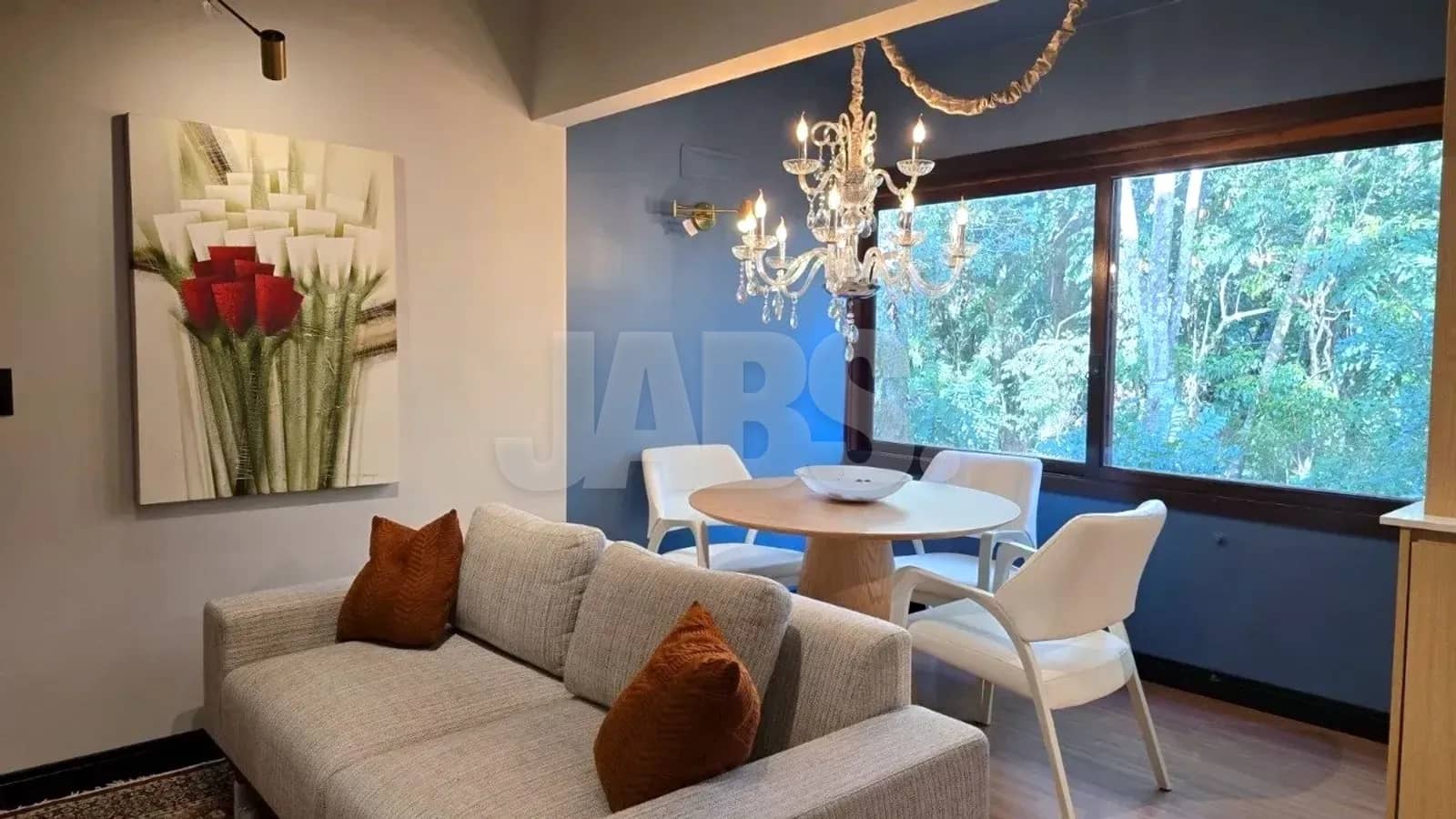 Sala de estar com sofá claro, almofadas terracota, lustre de cristal e janela para a mata