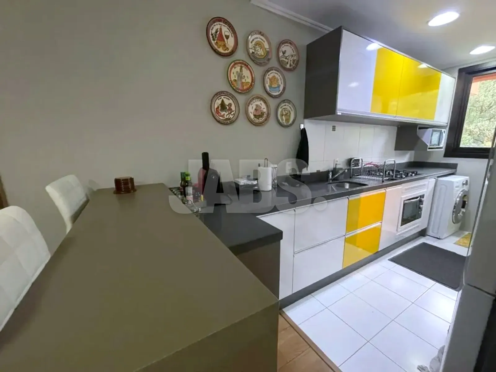 Cozinha com armários brancos e amarelos, bancada escura e pratos decorativos na parede