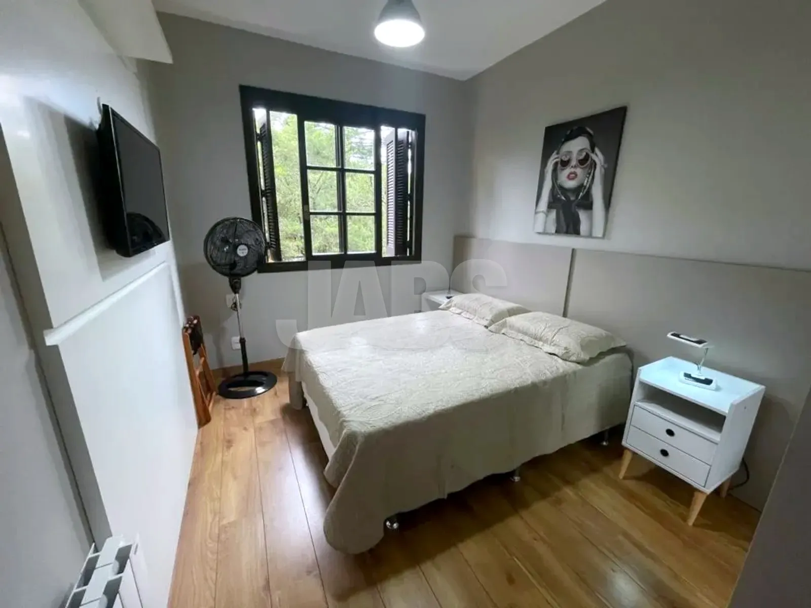 Suíte com cama de casal, criados-mudos brancos, TV e janela para o verde