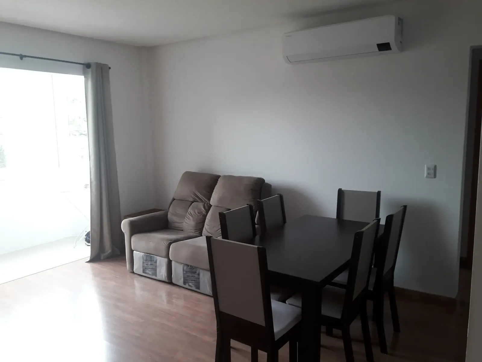 Sala com sofá de dois lugares, mesa de jantar para quatro, ar-condicionado split e porta de vidro para a sacada
