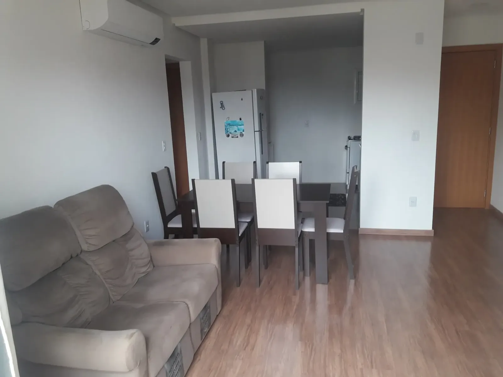 Sofá de tecido cinza na sala integrada à mesa de jantar, com a geladeira da cozinha americana ao fundo