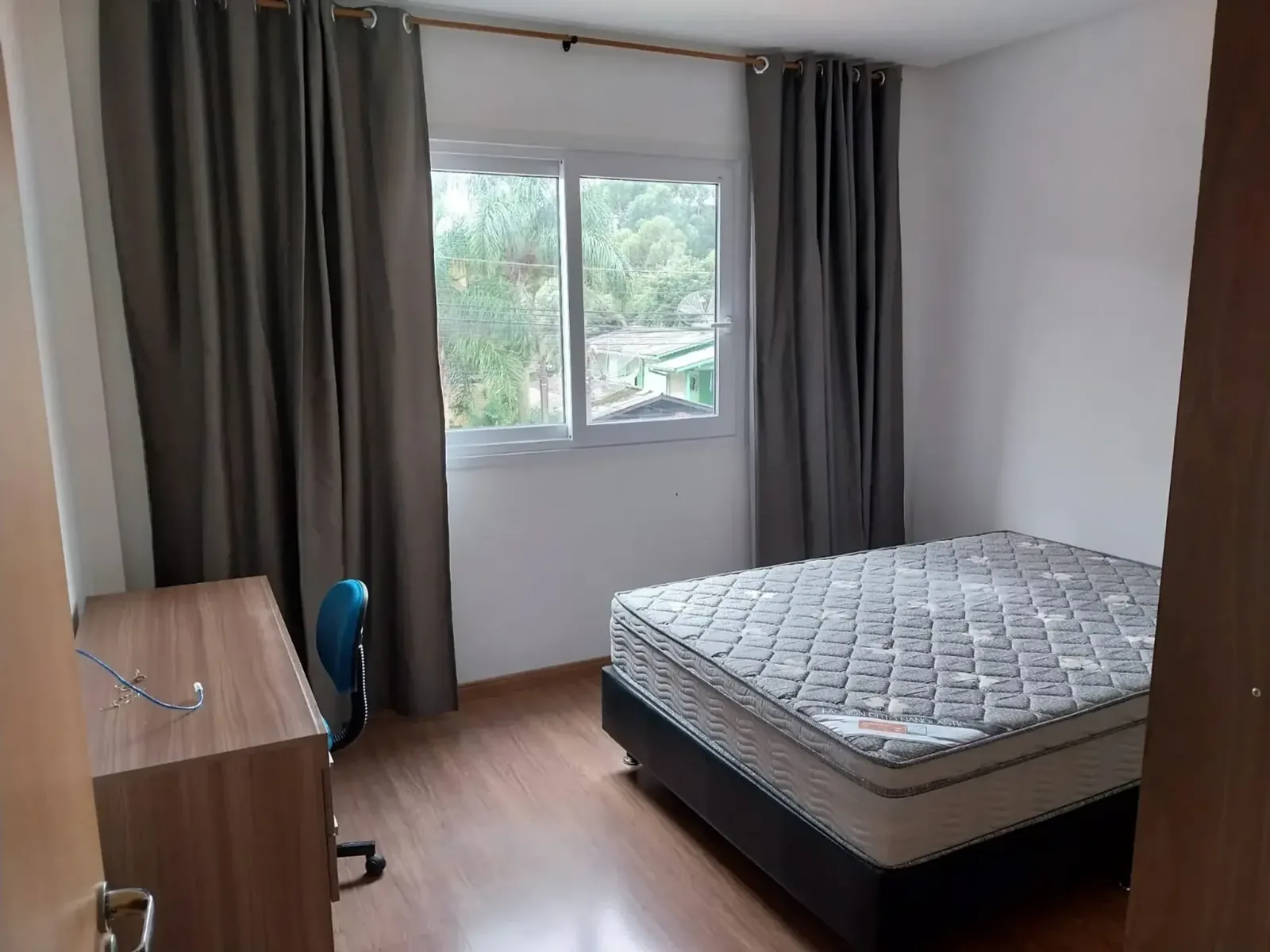 Dormitório com cama de casal, escrivaninha, cortinas e janela para as árvores