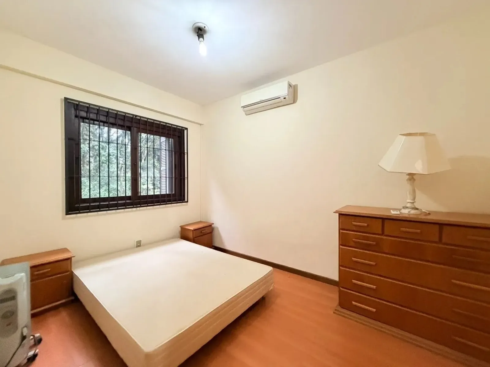 Dormitório com janela gradeada, mesa de cabeceira e cômoda de madeira