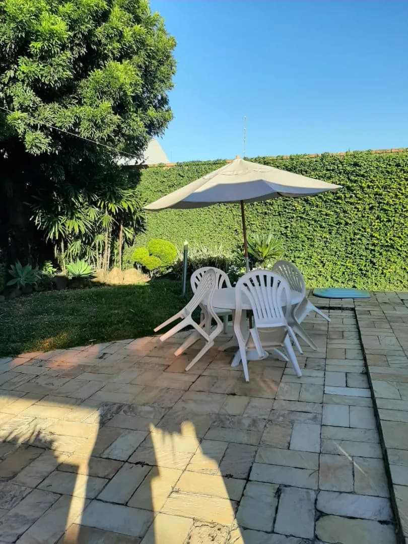 Pátio gramado com mesa, cadeiras e guarda-sol à sombra da cerca-viva.