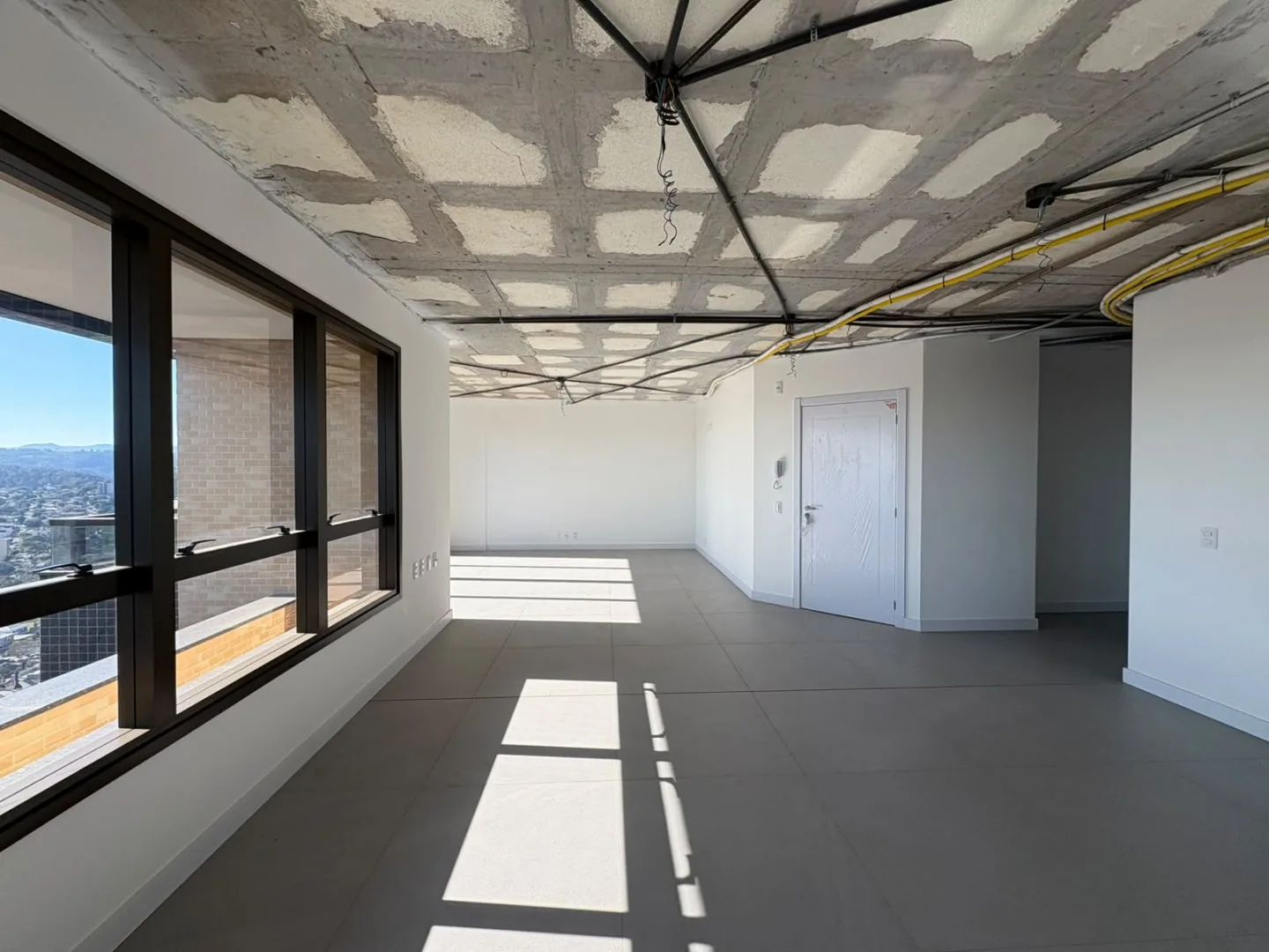 Ambiente em obra com janelas, porta branca e passagem para o corredor.