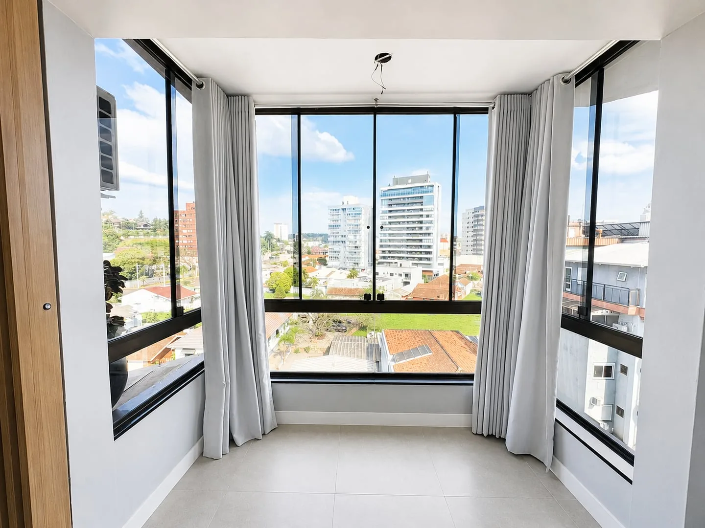 Ambiente vazio com janelas de canto, cortinas cinza e piso porcelanato claro.
