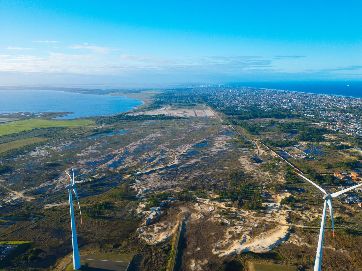Vista de drone com a lagoa à esquerda, o mar à direita e a obra do loteamento no meio, em julho de 2026