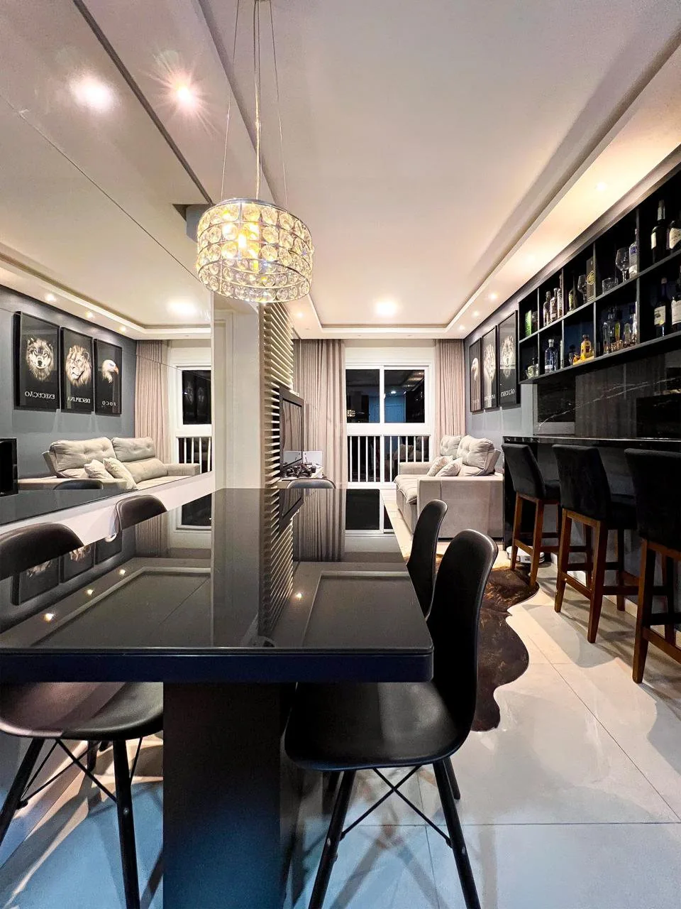 Cantinho bar com mesa e banquetas pretas, sob pendente de cristal, ao lado da sala.