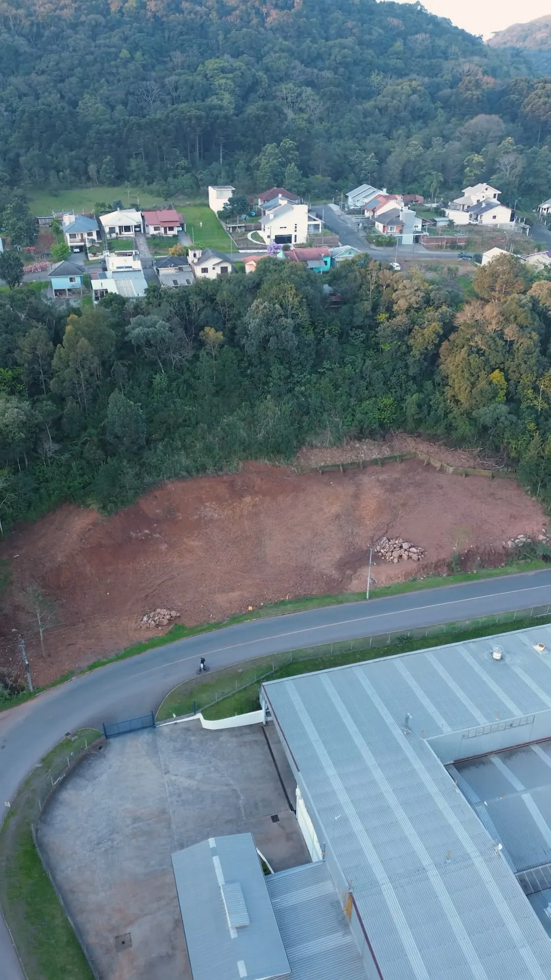 O terreno junto à rua, com as casas do bairro Piá no alto do morro e um pavilhão industrial do outro lado.