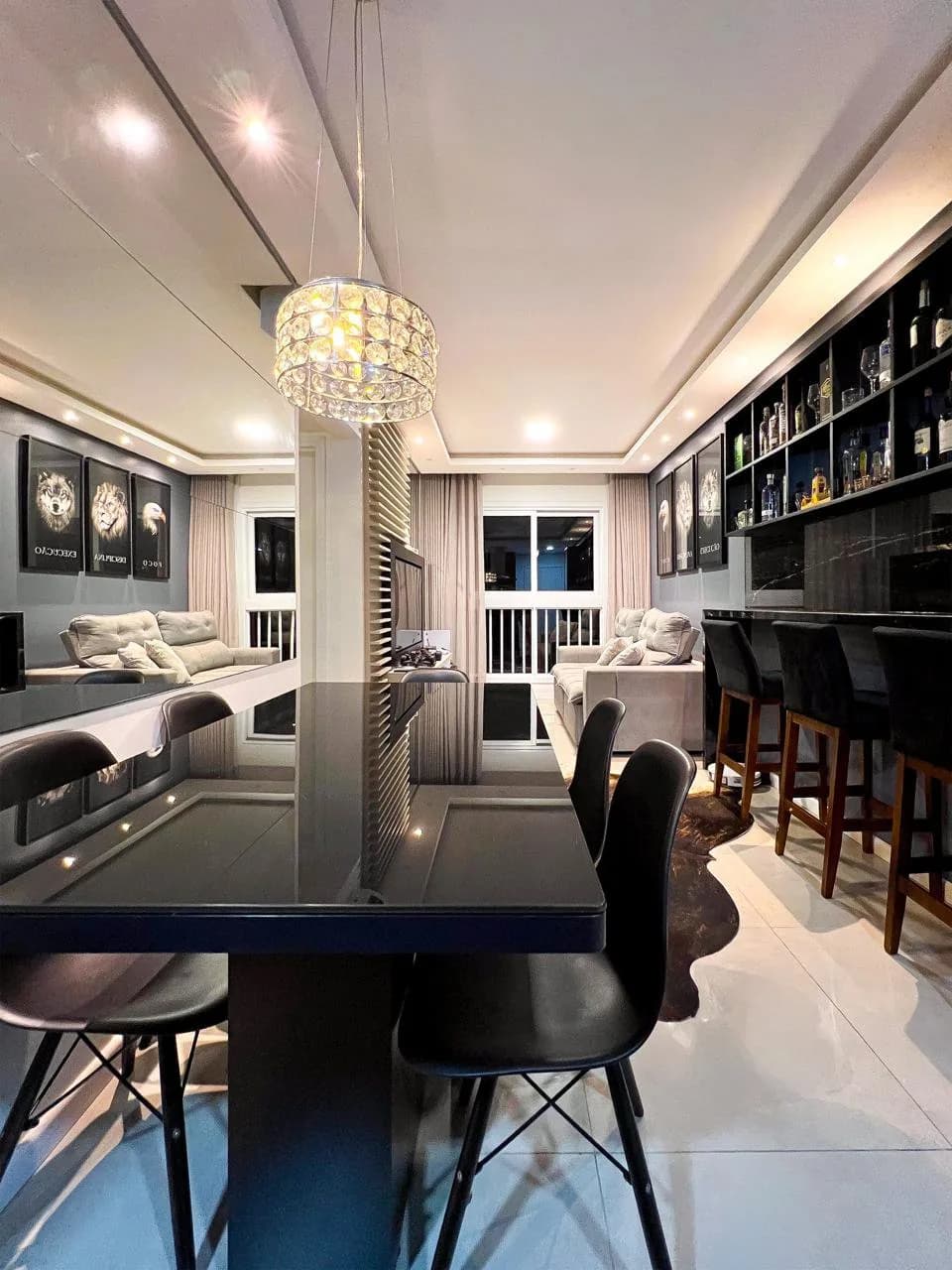 Cantinho bar com mesa e banquetas pretas, sob pendente de cristal, ao lado da sala.