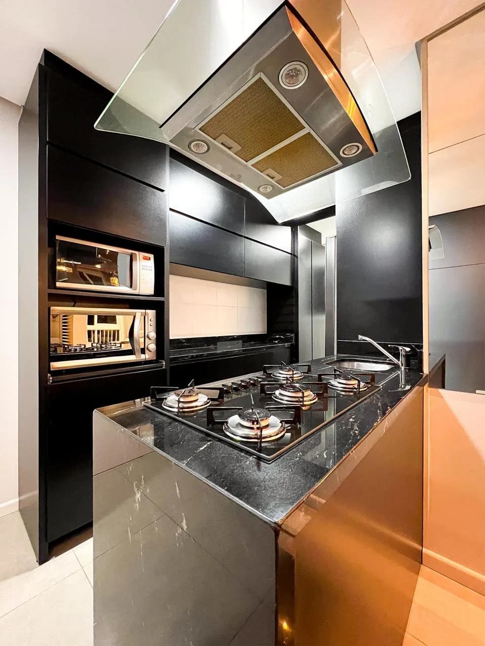 Cozinha planejada, com bancada preta, cooktop, coifa e acabamento em tom cobre.